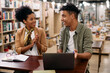 © Drazen - Happy African American student uses laptop and talks to his professor who is congratulating him on well done assignment in library.