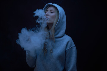 Wall Mural - Portrait of beautiful blonde girl in gray hoodie smokes vape isolated on black studio background, cloud of steam smoke, mini hookah