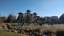 A Very Big Playground Castle For Children To Play. It Has Three Floors, Several Slides, Four Towers. Children Are Protected By Net. There Is Also A Stream In The Foreground Covered With Reed. Park Is 