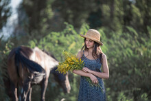 Young Woman In Blue Dress And Sun Hat Holding Yellow Flowers Standing Beside Brown Horse