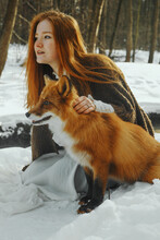 Young Woman With Red Hair Sitting On Snow Covered Ground With A Fox