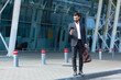 © Liubomir - stylish bearded business man with travel bag and mobile phone in hands walks on city street modern urban background. Businessman in a suit goes and uses a smartphone. tourist trip. caucasian manager