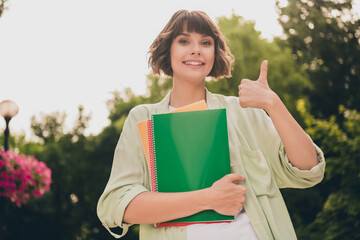 Poster - Portrait of attractive cheerful girl holding academic copybook showing thumbup advert on fresh air outdoors