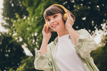 Wall Mural - Photo of charming pretty young lady dressed green shirt headphones walking smiling outdoors urban park