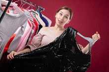 Young Woman Trying To Choose A Dress To Wear, Holding Black Clothes On A Hanger With A Hesitant Look, Trying To Make A Decision