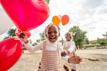 Positive Black Girls With Balloons Standing On Grass In Park