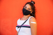 © loreanto - Portrait of young woman wearing protective face mask on orange wall after getting vaccine against Coronavirus Covid-19 infections. Millennial smile with medical plaster on arm - Copy space