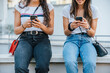 © loreanto - Detail of the hands of two young women sitting on a low wall while using the smartphone to surf social networks - Unrecognizable people - Millennial having fun together