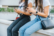 © loreanto - Detail of the hands of two young women sitting on a low wall while using the smartphone to surf social networks - Unrecognizable people - Millennial having fun together