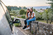 © loreanto - Portrait of two young women sitting on the low wall at sunset in a car trip among nature, behind them the city and vegetation - Millennials having fun together during an unforgettable vacation