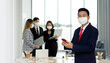 © chadchai - Asian businessman in a suit and face mask typing on mobile phone. The team is preparing for the meeting in the background. Everyone wears a mask. Concept of leadership.