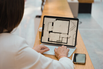 architect or interior designer works remotely. young freelance woman works with an engineering floor plan. brunette in a white jacket with a laptop in public space, drawing of apartment is on screen.