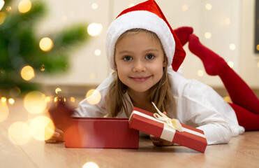 Little girl in red Santa Claus hat lies on floor with open box on background of christmas tree. Child with New Years gift looks at camera and smiles