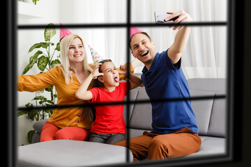 Wall Mural - Young big family celebrating Christmas enjoying dinner, view from outside through a window into a decorated living room with tree and candle lights, happy parents eating with three kids.