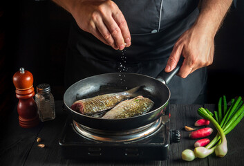 Professional chef prepares fresh fish in pan sprinkling salt with ingredients. Preparing to cook fish food. Working environment in the restaurant kitchen