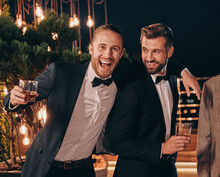 Two Handsome Men In Suits Drinking Whiskey And Smiling While Spending Time On Party