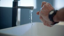 Caucasian man washes and disinfects his hands with with soap while a tap is running, he then starts to rinse his hands. He wears a watch and ring.