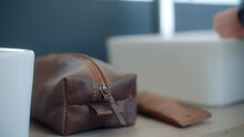 Caucasian male wearing a watch and ring zips closed a stylish leather toiletry bag in a modern bathroom.