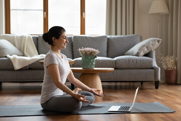 E yoga remotely, no stress, quarantine, mindfulness concept. In modern cozy living room at home young Indian woman wear sportswear sit on mat near laptop do meditation practice with on-line trainer