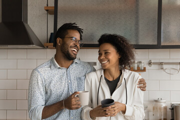 Laughing affectionate young African American family couple drinking morning coffee, standing in modern kitchen, talking speaking joking on weekend at home, spending leisure carefree time together.