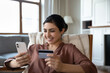 © fizkes - Happy Indian woman sit on armchair holds debit card and smart phone buying on internet. Purchasing goods, retail services online, remote comfort usage of modern technology, secure e-bank app concept