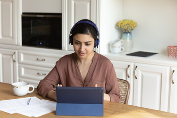 Canvas Print - Busy Indian woman sit at table listen audio course, colleague, tutor through headphones learns online use internet, videocall on tablet. Study, lead business negotiations via video call event concept