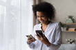 © fizkes - Smiling African American woman using two phones, standing at home, happy attractive young female looking at screens, browsing apps, synchronizing information on devices, setting up new smartphone