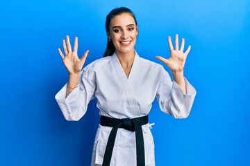 Wall Mural - Beautiful brunette young woman wearing karate fighter uniform with black belt showing and pointing up with fingers number ten while smiling confident and happy.