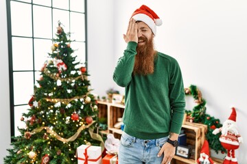 Canvas Print - Redhead man with long beard wearing christmas hat by christmas tree covering one eye with hand, confident smile on face and surprise emotion.