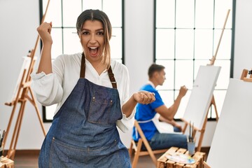 Canvas Print - Young hispanic couple at art studio screaming proud, celebrating victory and success very excited with raised arms