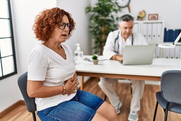 Sticker - Senior woman sitting at doctor appointment with hand on stomach because indigestion, painful illness feeling unwell. ache concept.