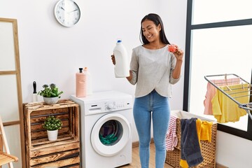 Wall Mural - Young latin woman holding detergent bottle and virus toy at laundry room