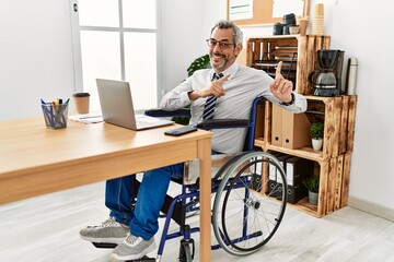 Canvas Print - Middle age hispanic man working at the office sitting on wheelchair smiling and looking at the camera pointing with two hands and fingers to the side.