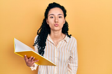 Young hispanic woman with curly hair holding book looking at the camera blowing a kiss on air being lovely and sexy. love expression.