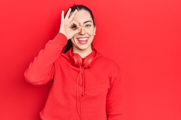 Poster - Beautiful woman with blue eyes wearing gym clothes and headphones smiling happy doing ok sign with hand on eye looking through fingers