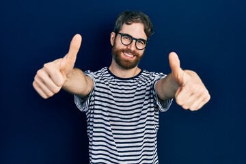 Caucasian man with beard wearing striped t shirt and glasses approving doing positive gesture with hand, thumbs up smiling and happy for success. winner gesture.