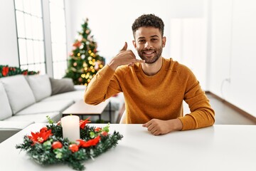 Wall Mural - Arab young man sitting on the table by christmas tree smiling doing phone gesture with hand and fingers like talking on the telephone. communicating concepts.