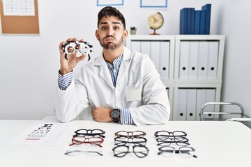 Sticker - Young optician man holding optometry glasses puffing cheeks with funny face. mouth inflated with air, crazy expression.