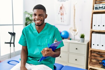 Canvas Print - Young african american man working at pain recovery clinic winking looking at the camera with sexy expression, cheerful and happy face.