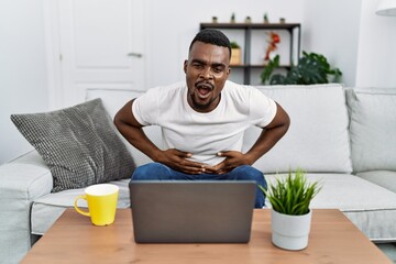 Wall Mural - Young african man using laptop at home with hand on stomach because indigestion, painful illness feeling unwell. ache concept.