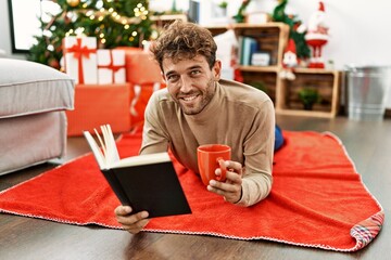 Poster - Young hispanic man reading book and drinking coffee lying by christmas tree at home