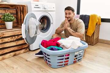 Wall Mural - Young handsome man putting dirty laundry into washing machine praying with hands together asking for forgiveness smiling confident.