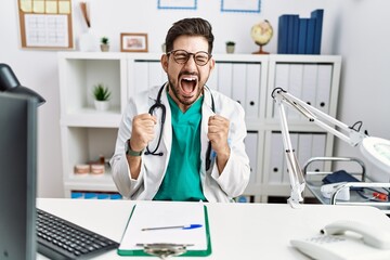 Poster - Young man with beard wearing doctor uniform and stethoscope at the clinic excited for success with arms raised and eyes closed celebrating victory smiling. winner concept.