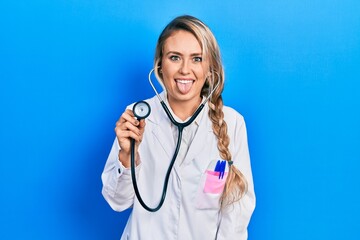 Sticker - Beautiful young blonde doctor woman holding stethoscope sticking tongue out happy with funny expression. emotion concept.