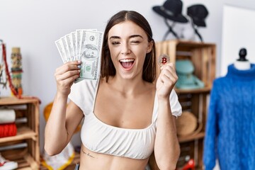 Poster - Young hispanic customer woman holding dollars banknotes and bitcoin winking looking at the camera with sexy expression, cheerful and happy face.