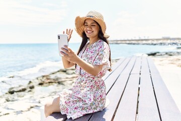 Poster - Young latin girl wearing summer hat making selfie by the smartphone sitting on the bench at the beach.