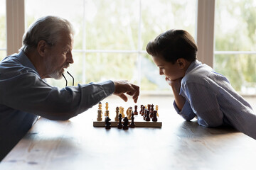 Your move grandpa. Aged grandfather retiree moving figure on chessboard while preteen grandson think how to win in game. Elderly granddad teaching little boy grandkid to play chess share experience