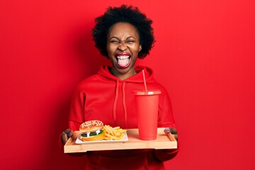 Wall Mural - Young african american woman eating a tasty classic burger with fries and soda sticking tongue out happy with funny expression.