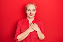 Young Blonde Woman Wearing Casual Red Shirt Smiling With Hands On Chest With Closed Eyes And Grateful Gesture On Face. Health Concept.