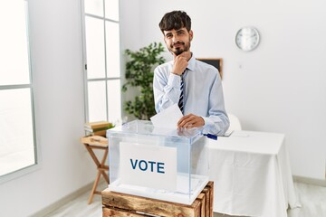 Poster - Hispanic man with beard voting putting envelop in ballot box looking confident at the camera smiling with crossed arms and hand raised on chin. thinking positive.